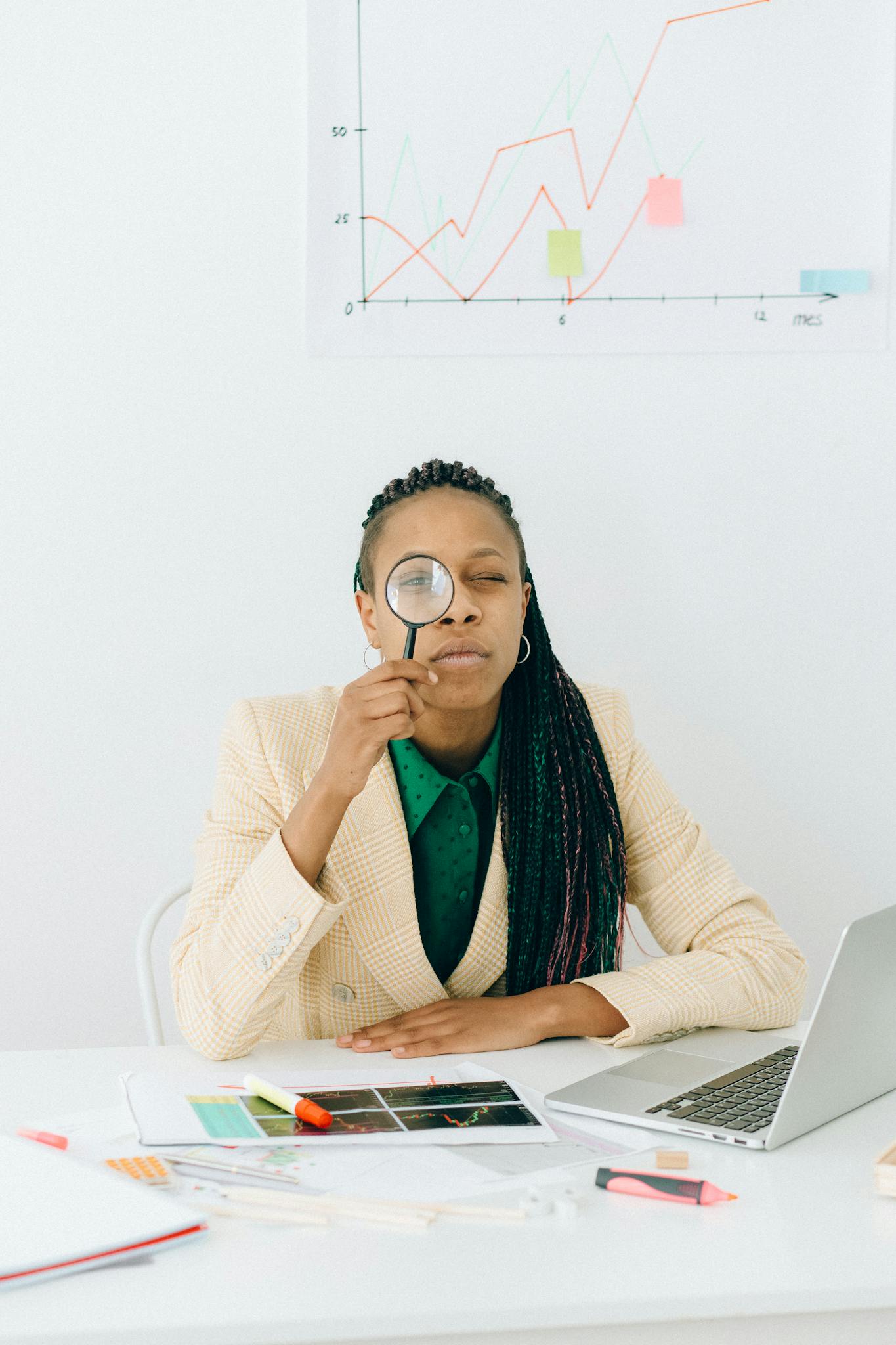 Woman examining data charts with a magnifying glass at a desk with a laptop, showcasing analytical skills.